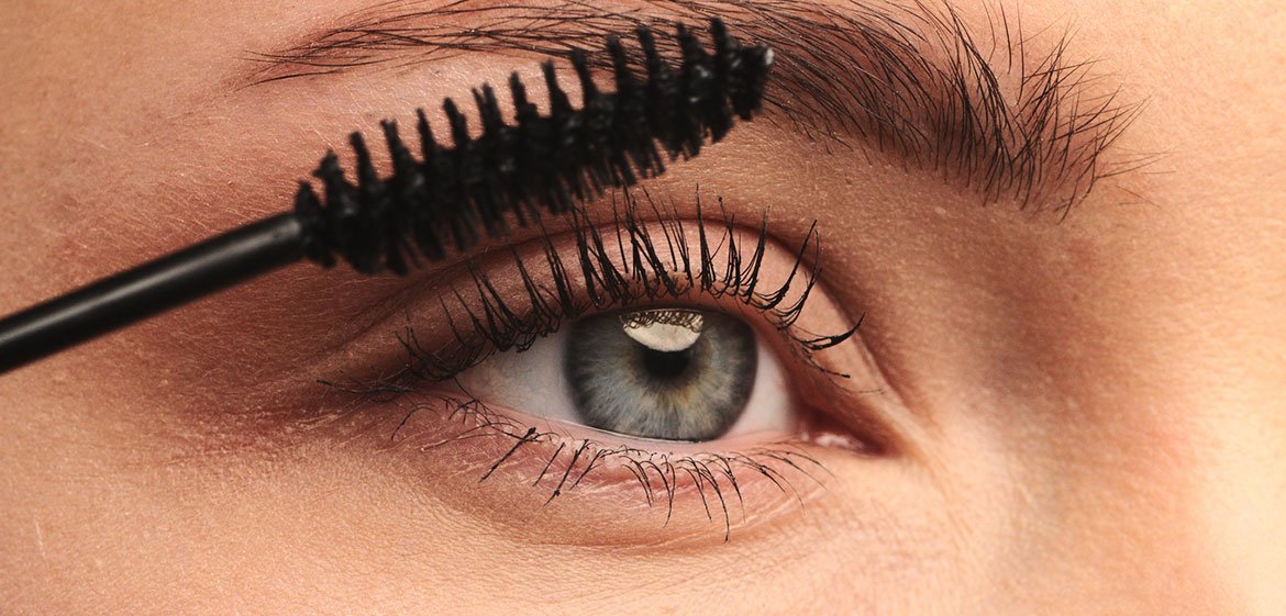 Nahaufnahme eines hellblauen Frauenauges, dessen Wimpern von einer Mascara-Bürste schwarz geschminkt werden.